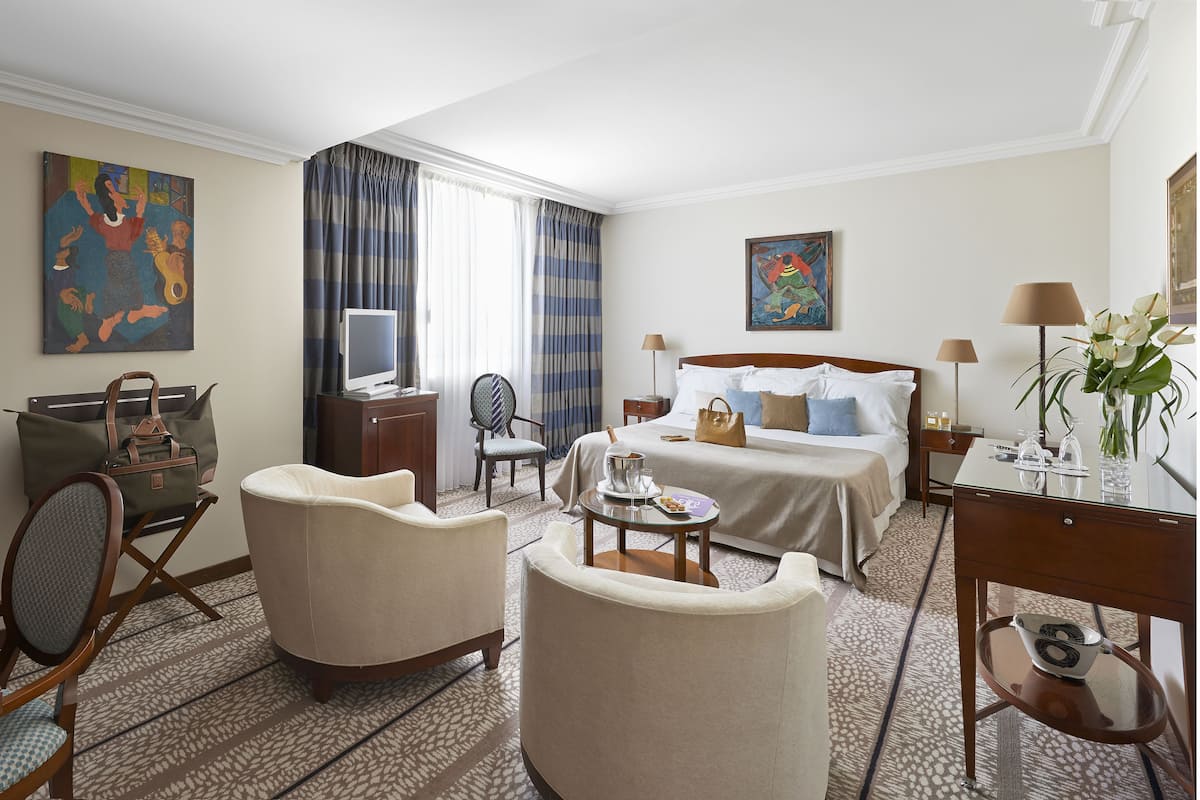 Habitación de hotel con cama, dos sillones, escritorio, televisor y obras de arte en las paredes.