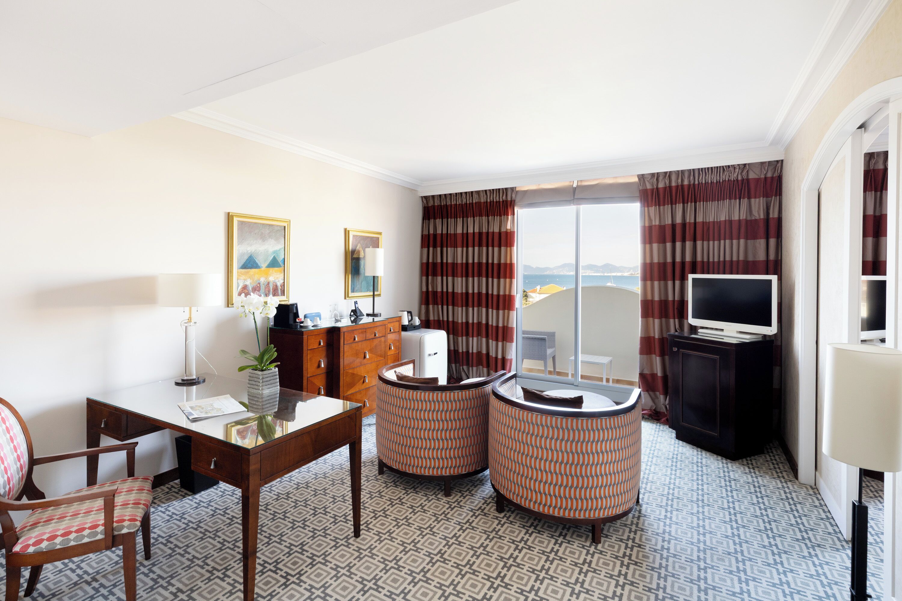 A hotel room with a dining table, two armchairs, a TV, and a view of the sea.