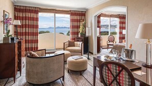 A hotel room with a large window, a sofa, a chair, a dining table, and a view of the sea. - Hotel Juana (Antibes)