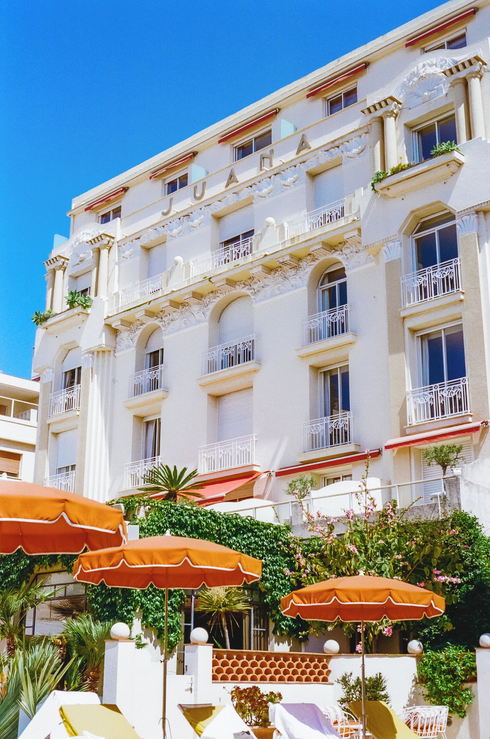 Un hotel de varios pisos con balcones, mobiliario exterior con sombrillas y abundante vegetación.