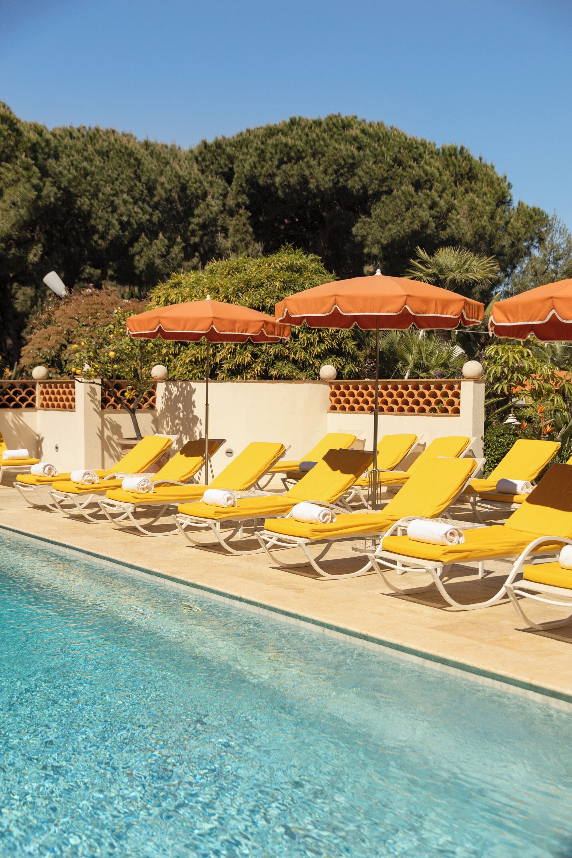 A poolside area with sun loungers and umbrellas, surrounded by a wall with a lattice pattern and greenery.