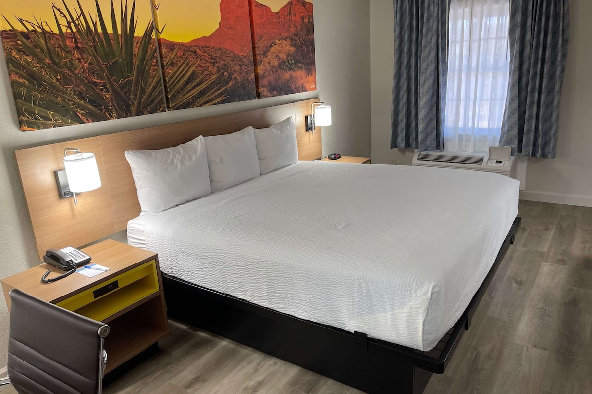 Ein Hotelzimmer mit einem großen Bett, einem Nachttisch aus Holz mit Telefon und Lampe, einem kleinen Tisch mit Lampe sowie einem Gemälde von Wüstenpflanzen an der Wand.