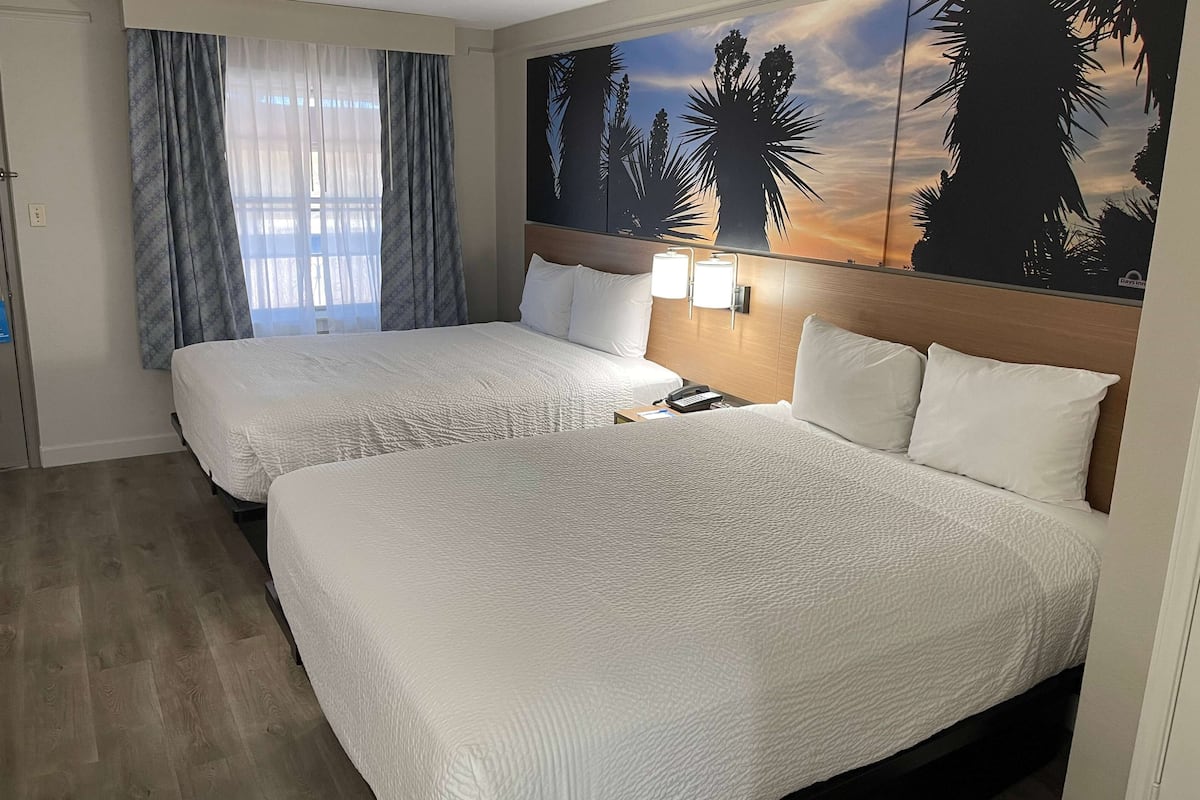 Ein Hotelzimmer mit zwei Betten, einem großen Fenster mit Vorhängen und einem Sonnenuntergangsbild an der Wand.