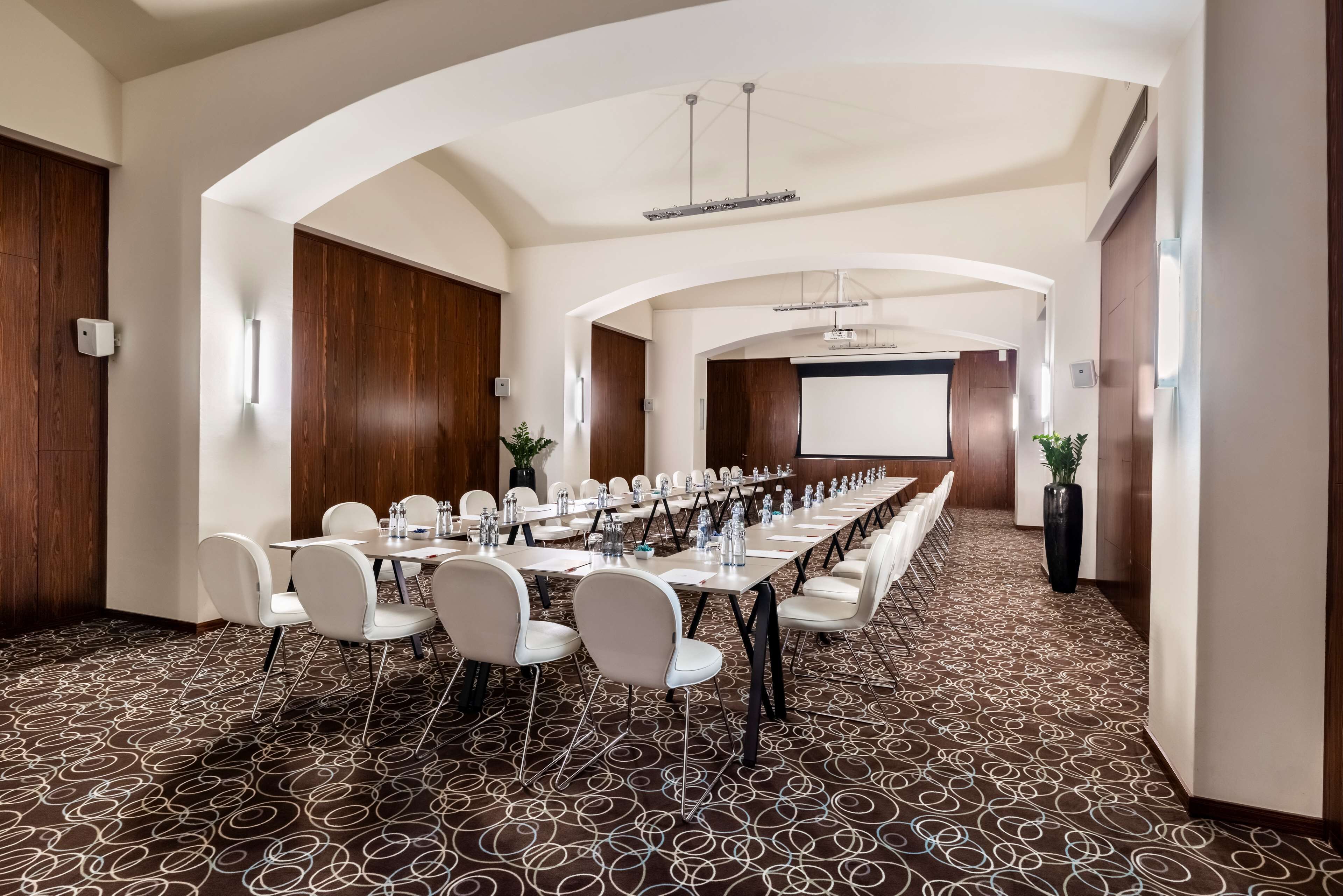 Uma sala de conferências com uma mesa comprida, cadeiras brancas e uma tela de projetor.