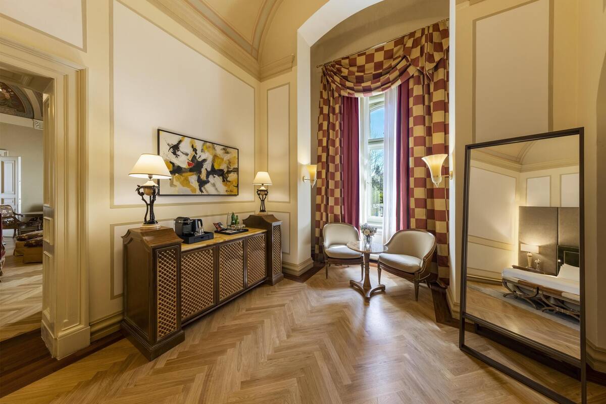 Une chambre d’hôtel avec un parquet en chevrons, un rideau à carreaux, un tableau, une console, deux chaises et un grand miroir.
