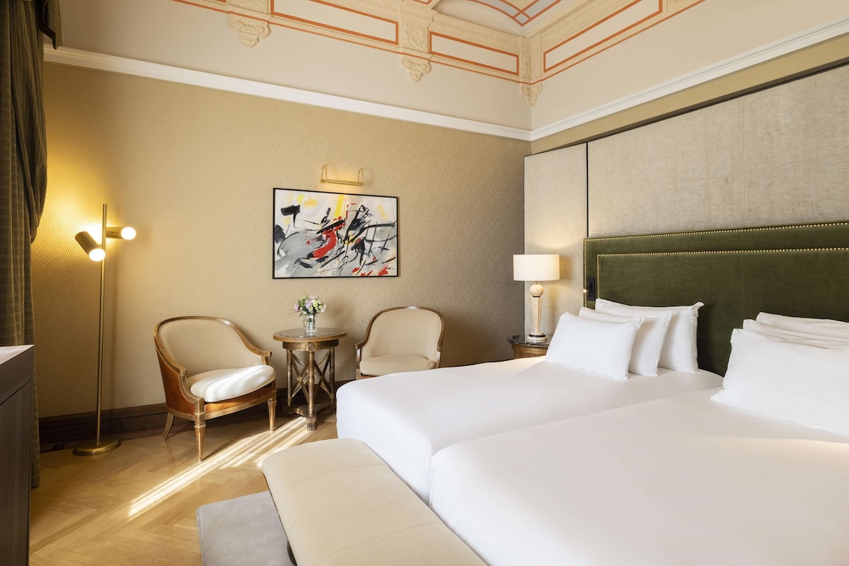 Une chambre d’hôtel avec un grand lit, deux chaises, une petite table avec un vase et un tableau au mur.