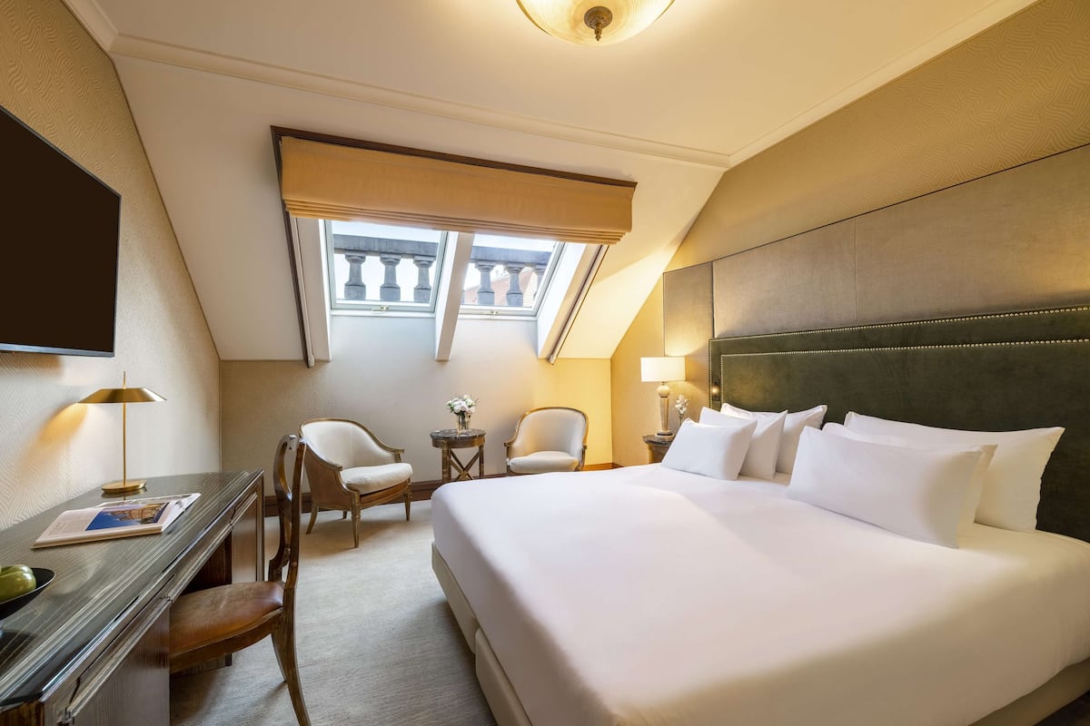 Une chambre d’hôtel avec un grand lit, un bureau avec une lampe, deux chaises, une petite table avec des fleurs et une fenêtre donnant sur des immeubles.