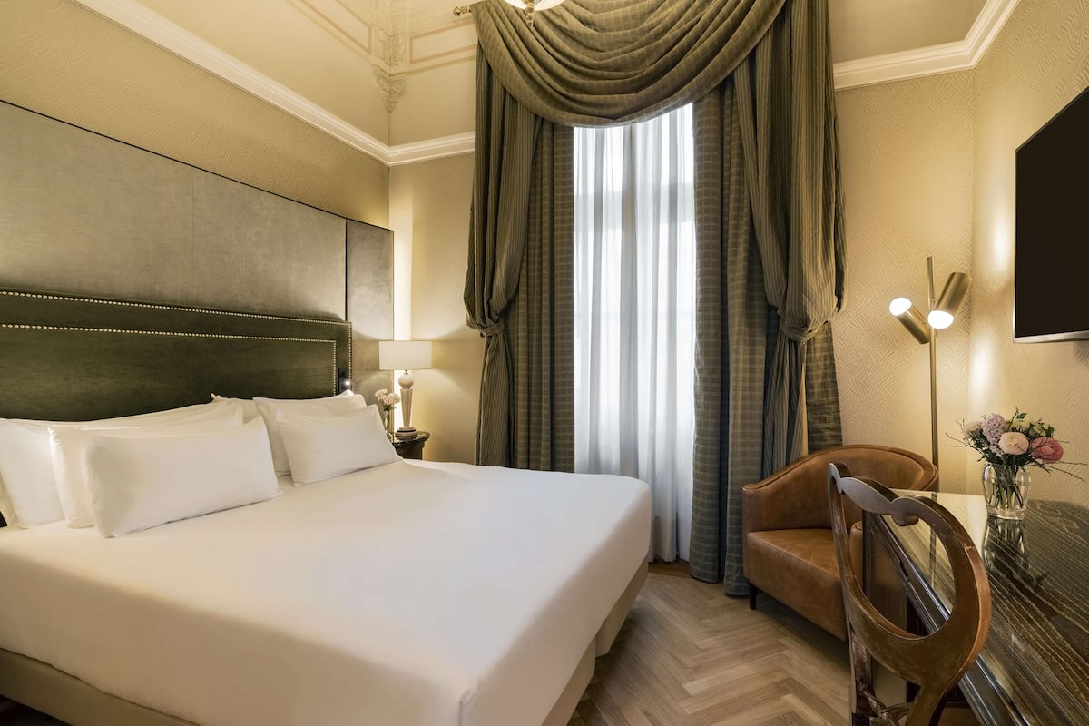 Une chambre d’hôtel avec un grand lit, une chaise en bois, un bureau avec une lampe et une vue sur l’extérieur à travers des rideaux.