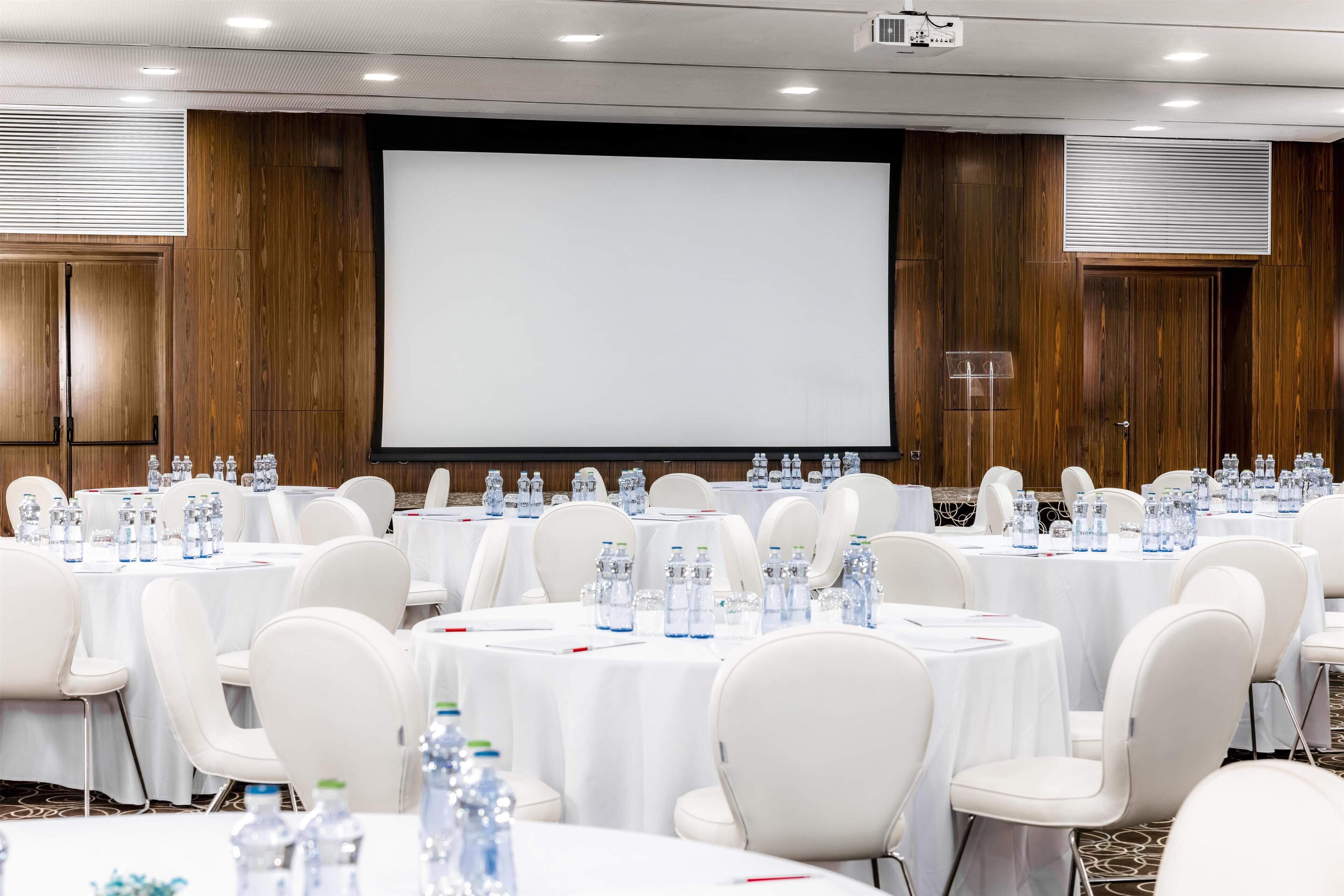 Uma sala de conferências com mesas redondas, cadeiras brancas e uma tela grande.