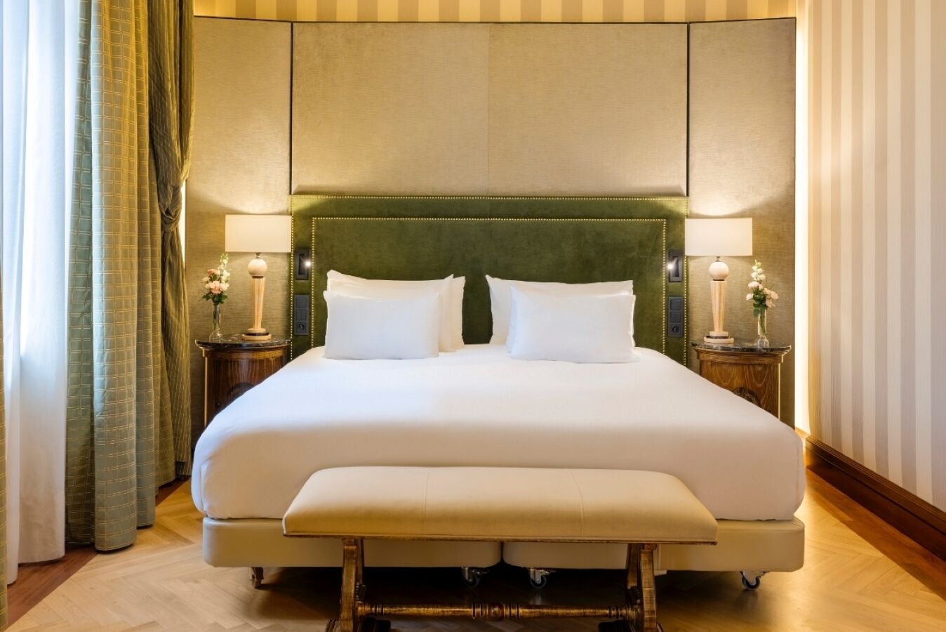 Uma cama grande com cabeceira estofada verde, duas mesas de cabeceira com abajures e um banco ao pé da cama.
