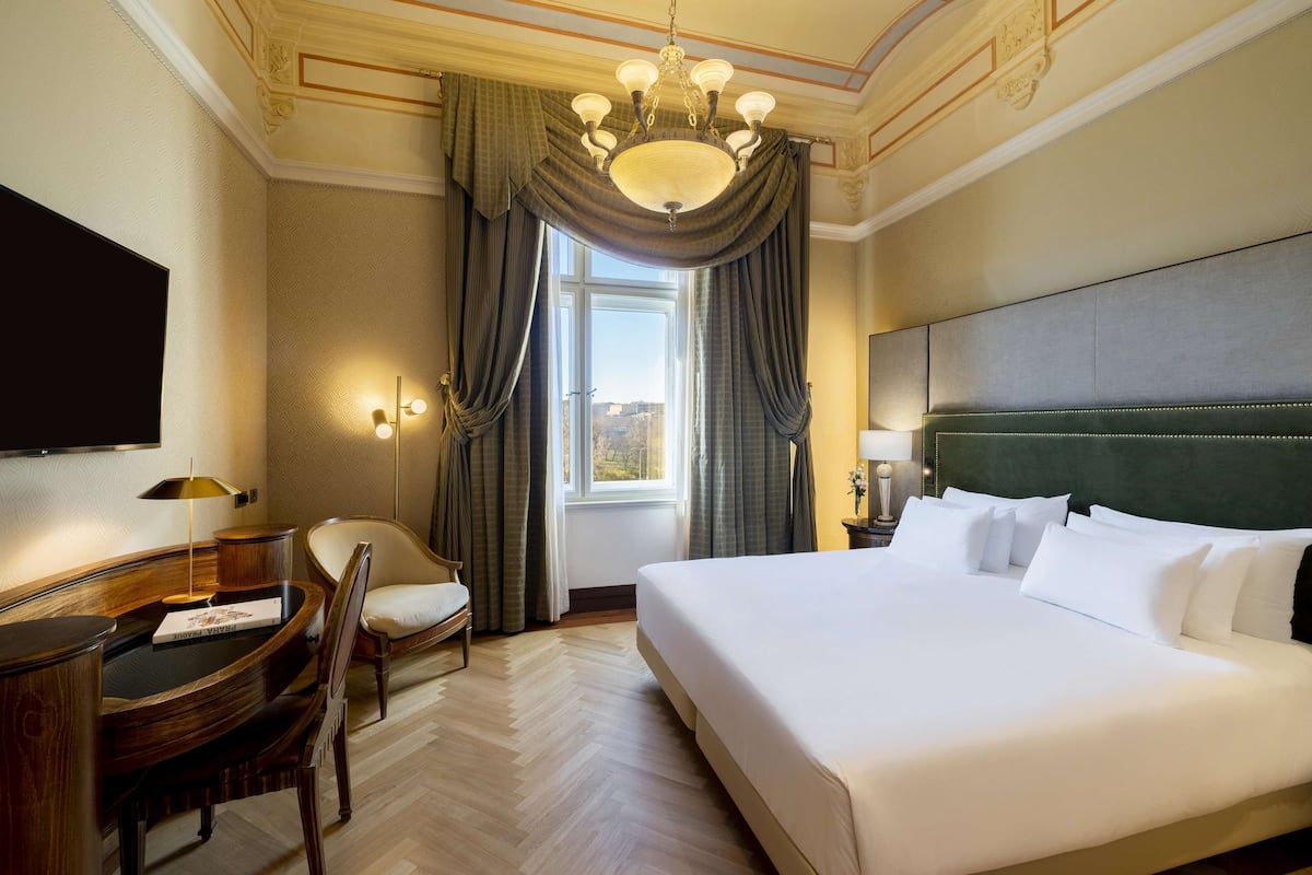 Une chambre d’hôtel avec un grand lit, un bureau avec une chaise, un téléviseur à écran plat, une fenêtre avec des rideaux et un lustre.