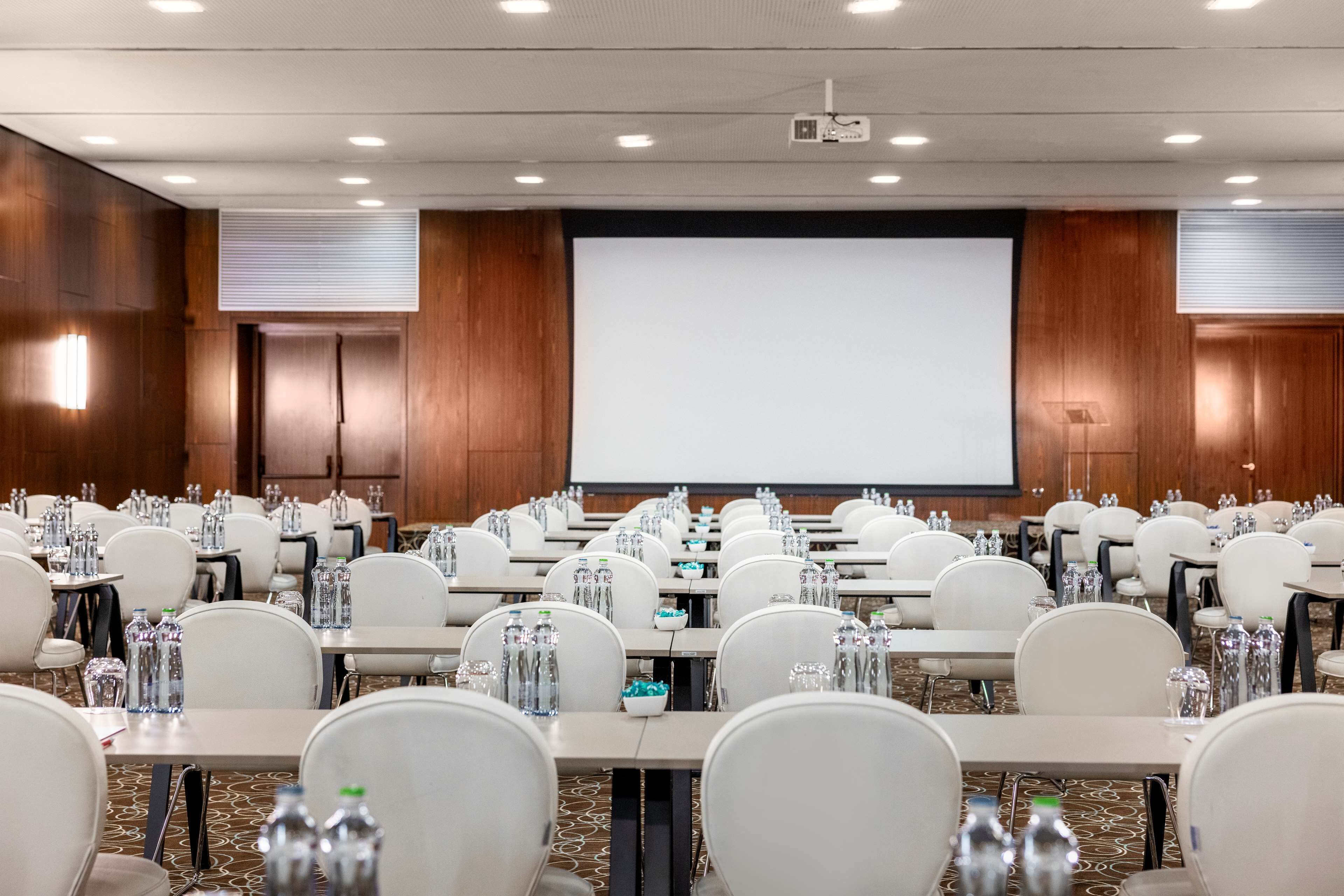 A conference room with rows of tables and chairs, equipped with water bottles and a projector screen.