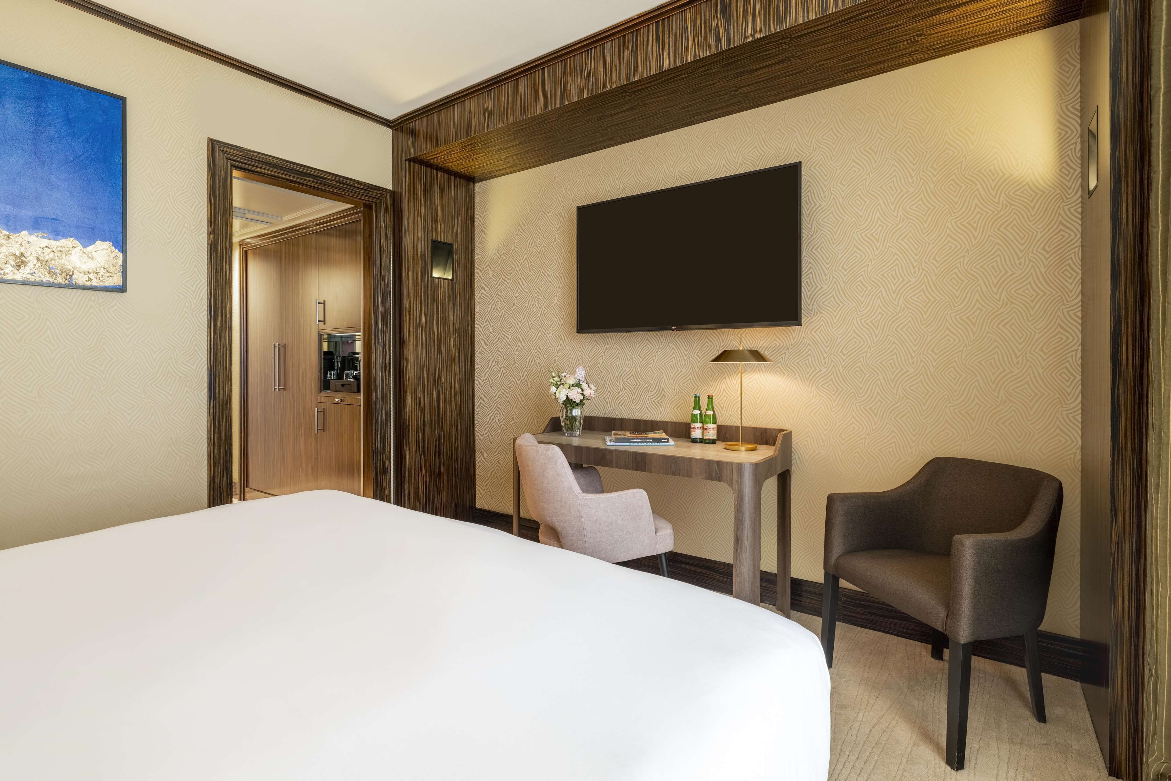 Quarto de hotel com uma cama grande, uma escrivaninha com cadeira, uma TV de tela plana e uma mesinha com um vaso e garrafas.