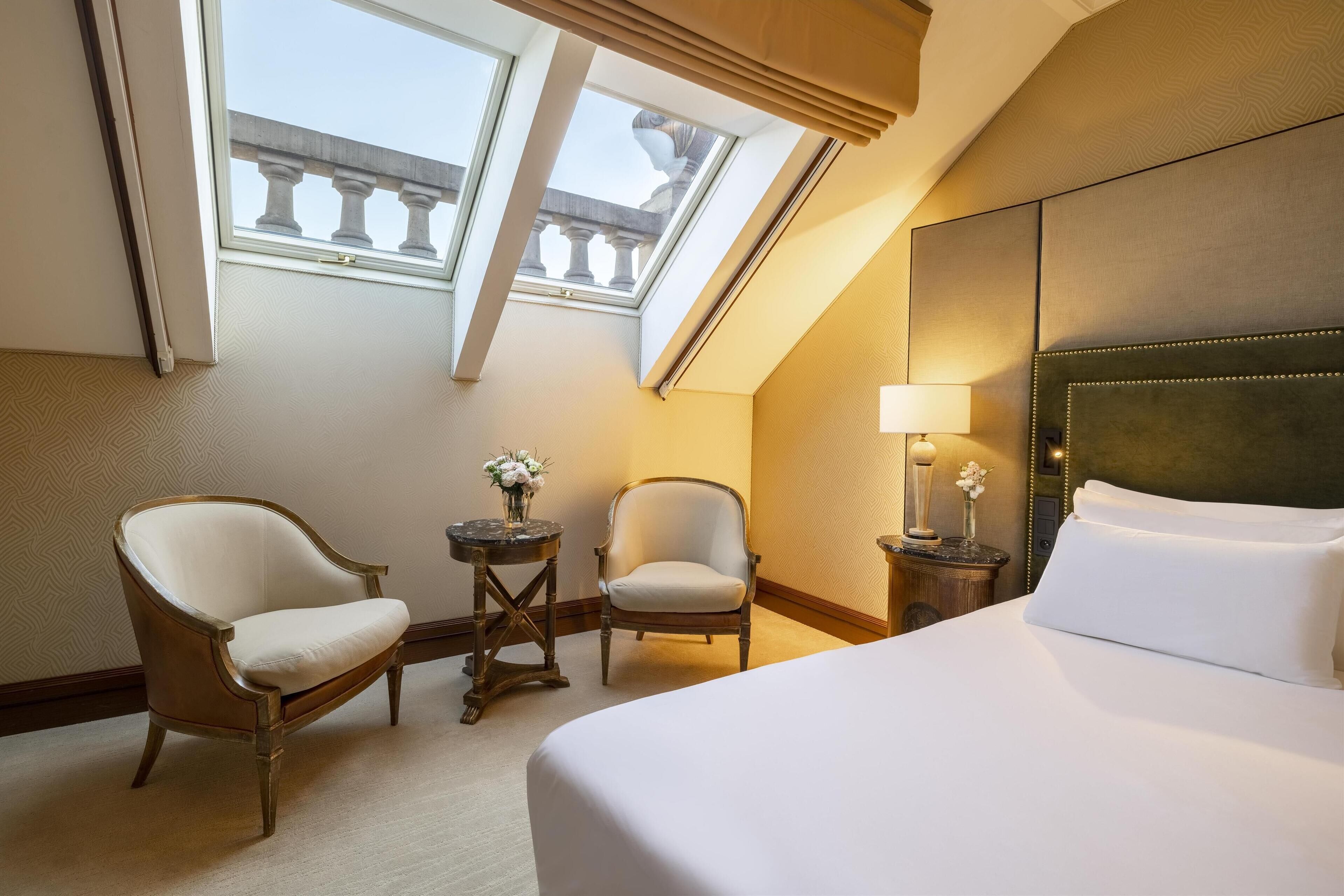 Camera d'albergo con un letto grande, due poltrone, un tavolino con una lampada e vista su un balcone attraverso degli skylight.