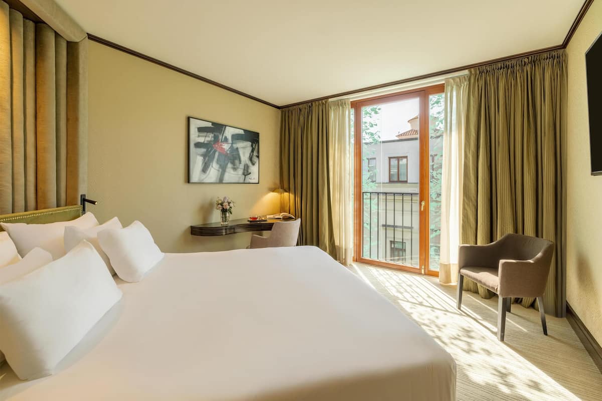 Une chambre d’hôtel avec un grand lit, un bureau avec une lampe, une chaise et une vue sur l’extérieur grâce à une porte vitrée.
