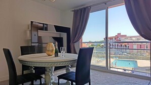Une chambre d’hôtel avec une table de salle à manger ronde, deux chaises, un vase et une bouteille de champagne. Une grande fenêtre offre une vue sur une piscine et des immeubles.