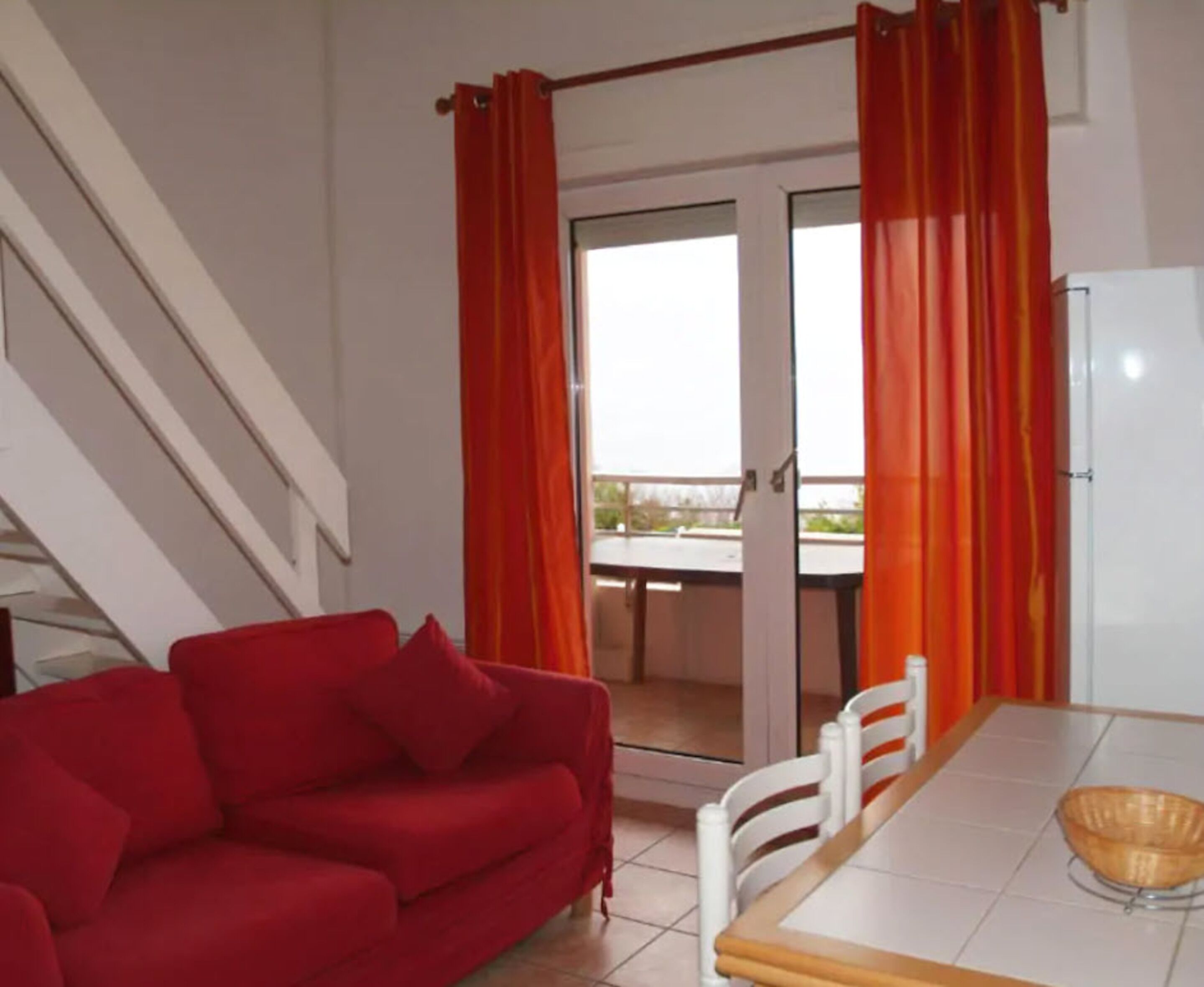 Un canapé rouge, une table à manger avec des chaises et un réfrigérateur dans une pièce donnant sur un balcon.