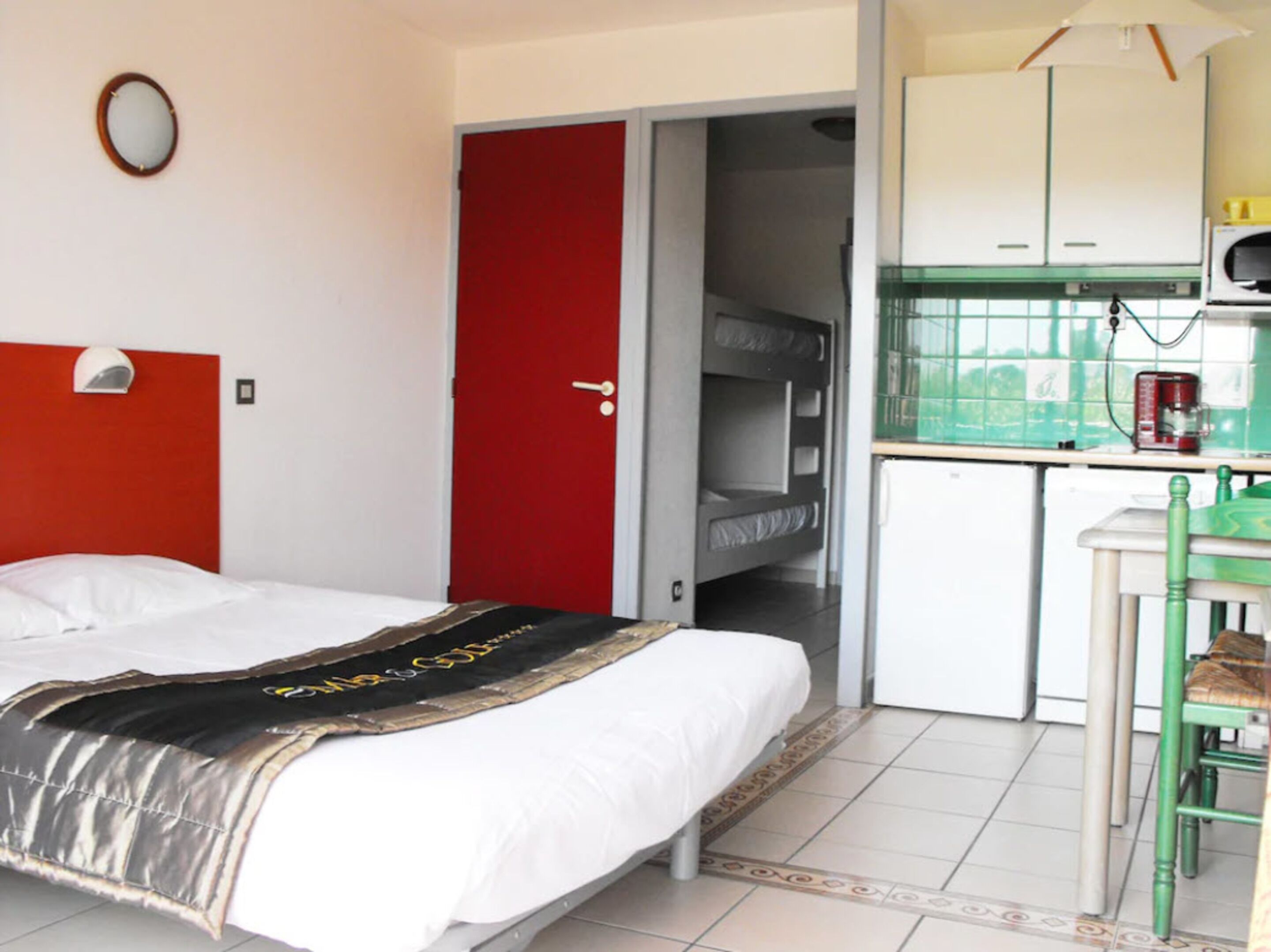 A hotel room with a bed, a red door, a kitchenette, and a small table with a chair.