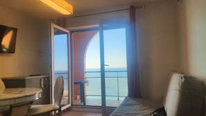 A room with a bed, a chair, a small table, and a view of the sea through a glass door.