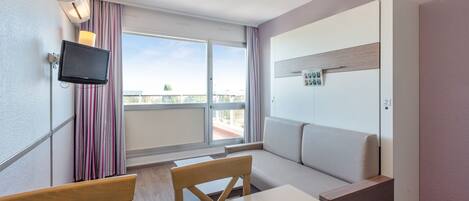 A modern room with a white sofa, a dining table with chairs, a flat-screen TV mounted on the wall, and a window with curtains.