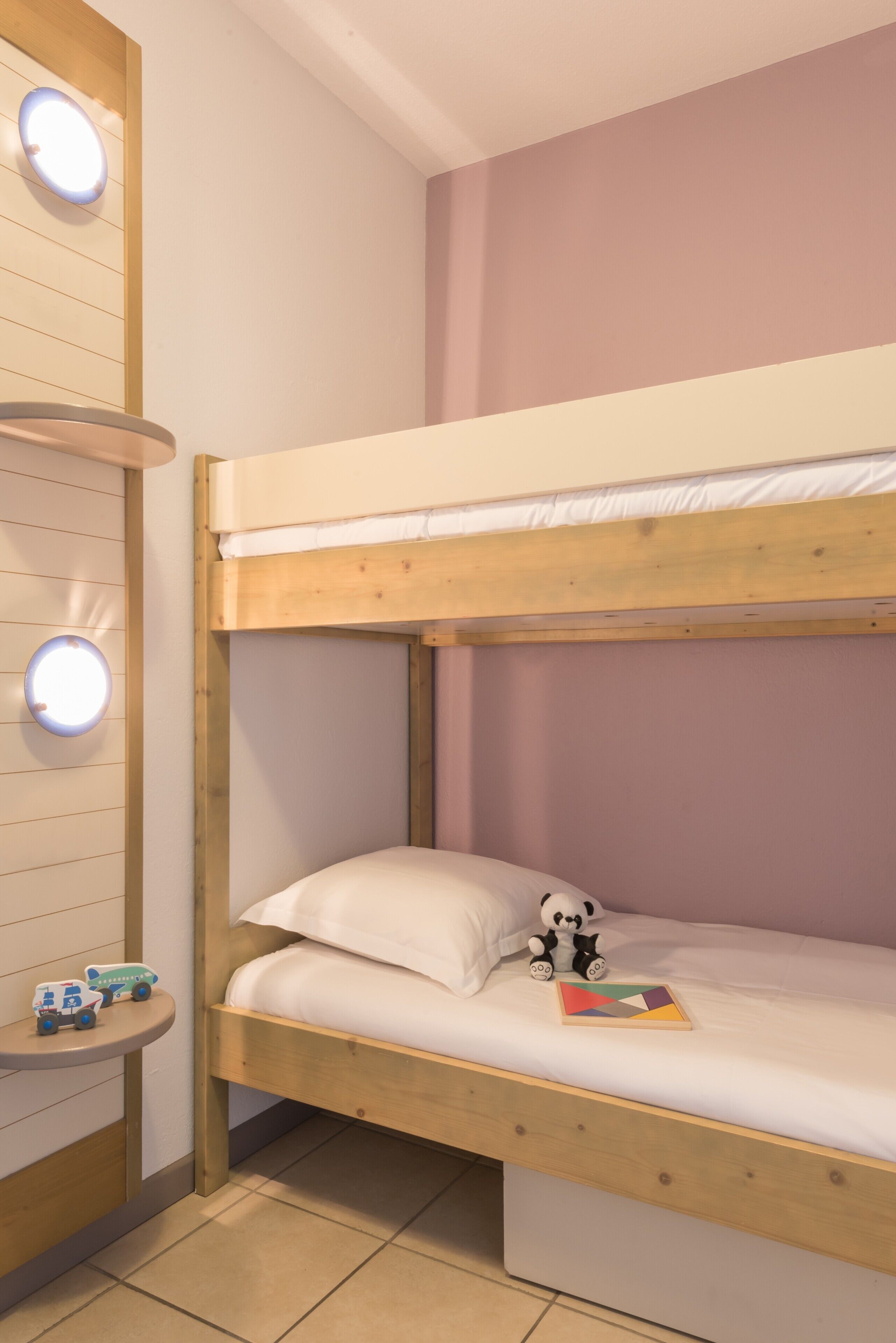 Un lit superposé avec une petite table et des jouets, un luminaire rond et un mur uni.