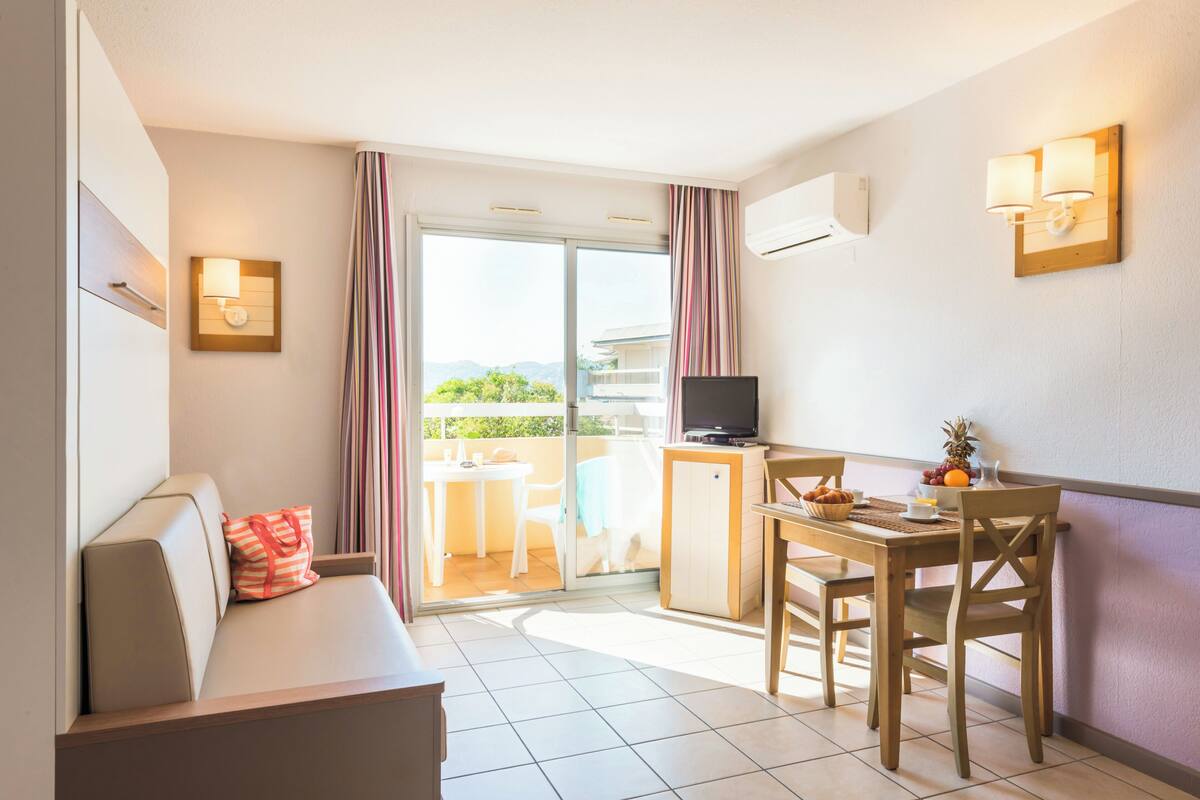 Ein Hotelzimmer mit einer Couch, einem Esstisch mit Stühlen, einem Fernseher und einem Balkon mit Vorhängen.