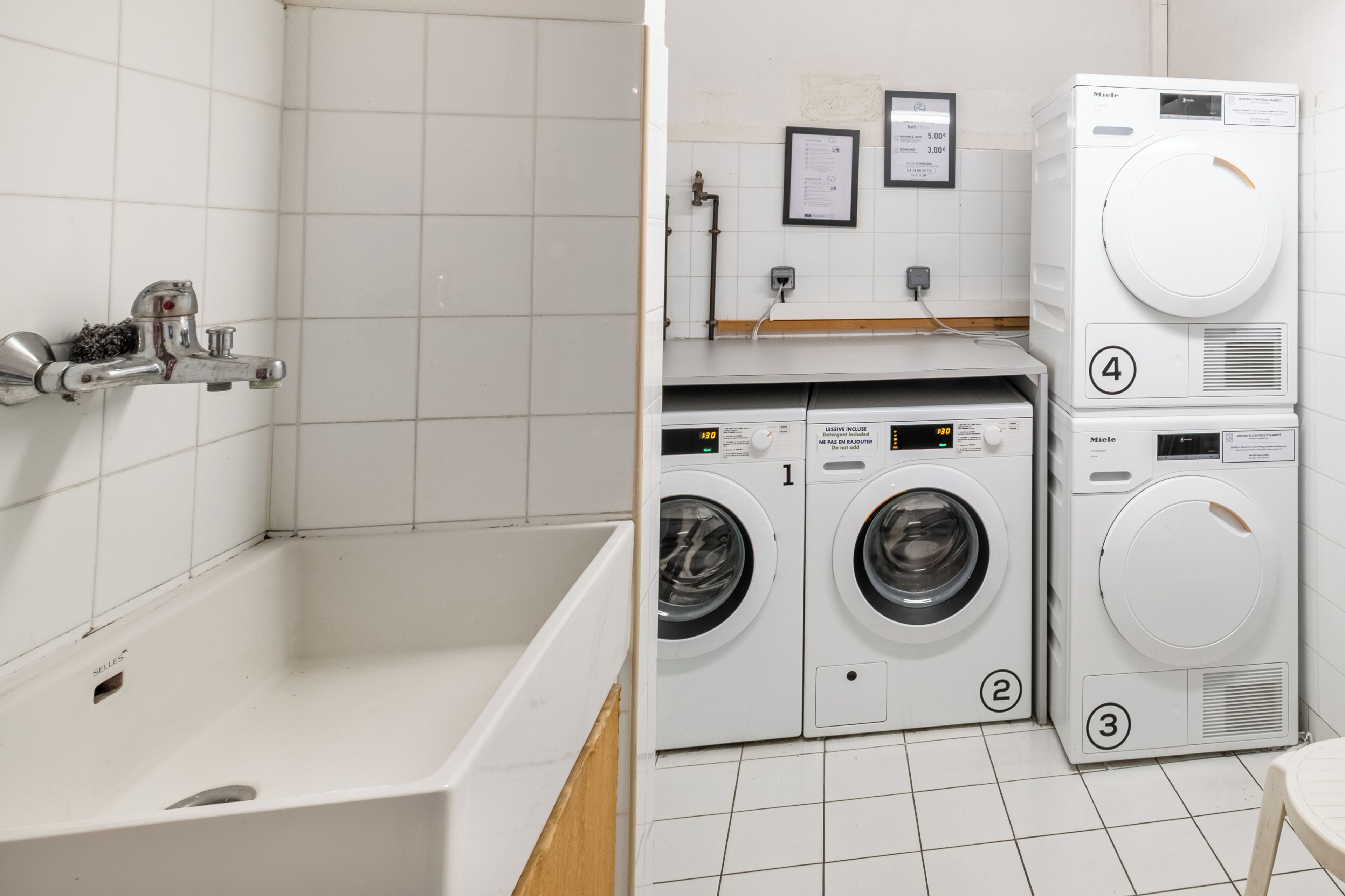 A laundry area with four stacked washing machines, a wall-mounted faucet, and a white tiled floor.