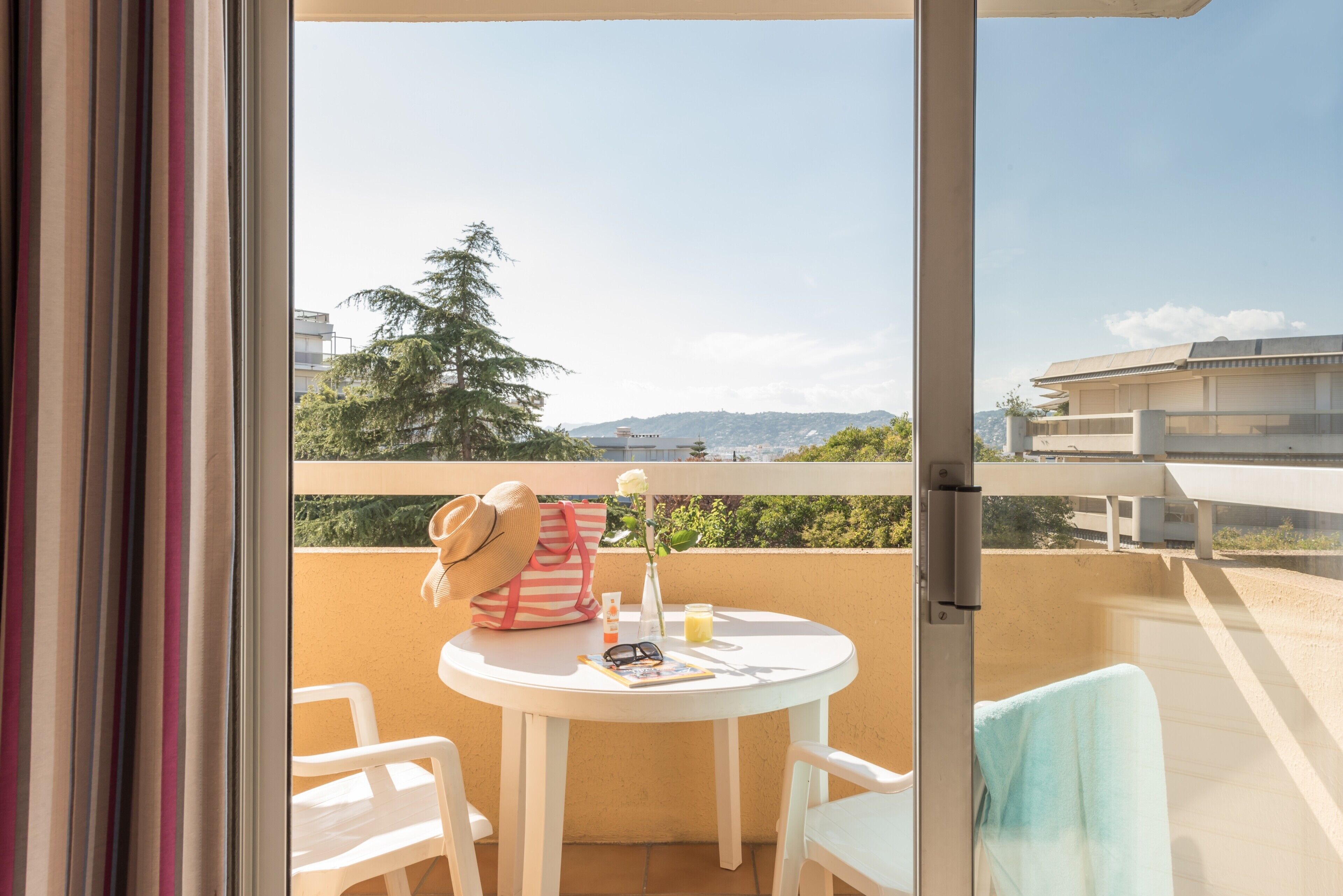 Ein Balkon mit Tisch, Stühlen, einem Hut, einer Tasche, einer Vase mit Blume und einem Glas Saft.