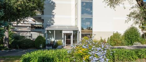 A modern building with a glass facade, a covered entrance, and a well-maintained garden with various plants and flowers.