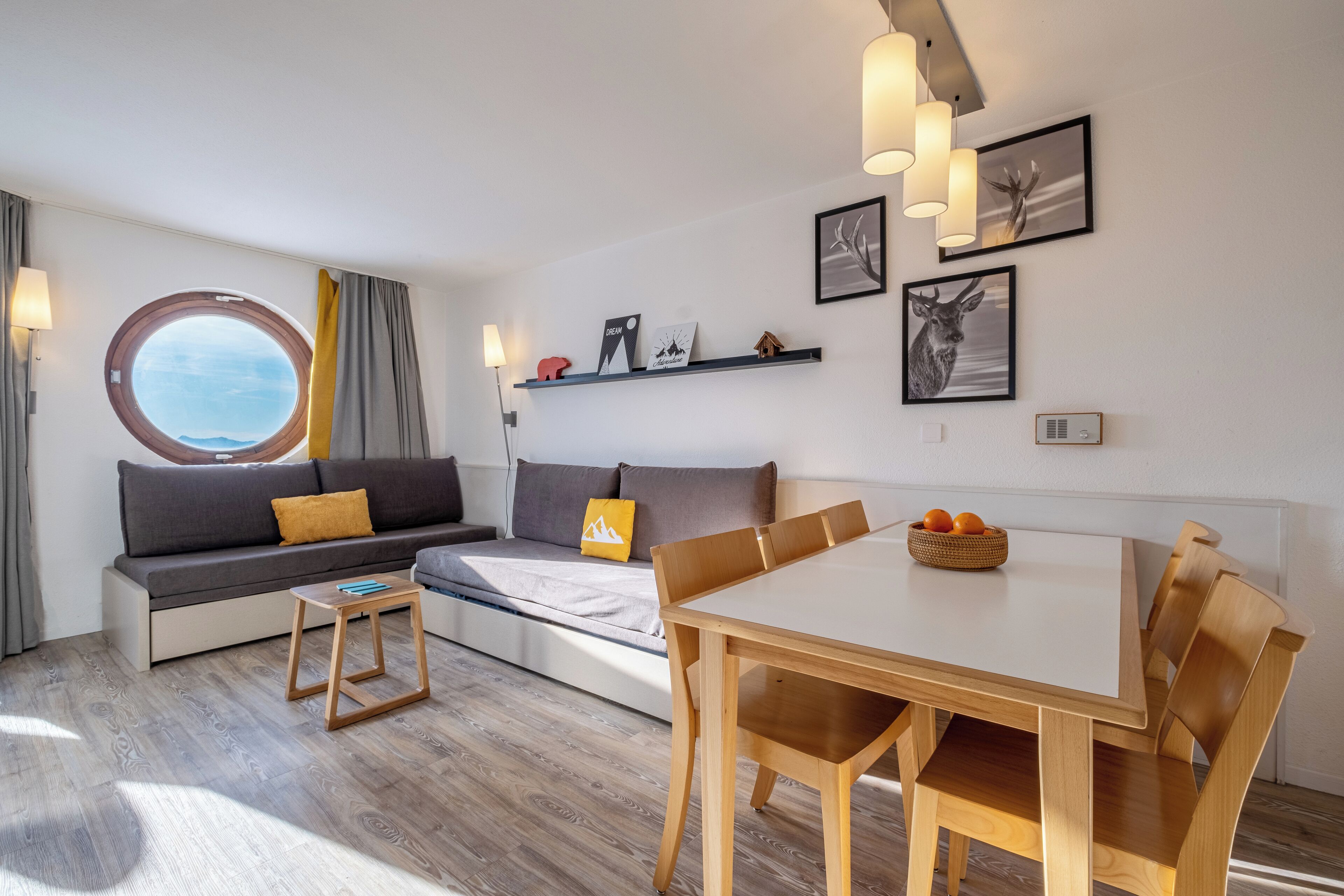 Un salon moderne avec un canapé, une fenêtre ronde, une table à manger avec des chaises et des étagères murales ornées de tableaux.