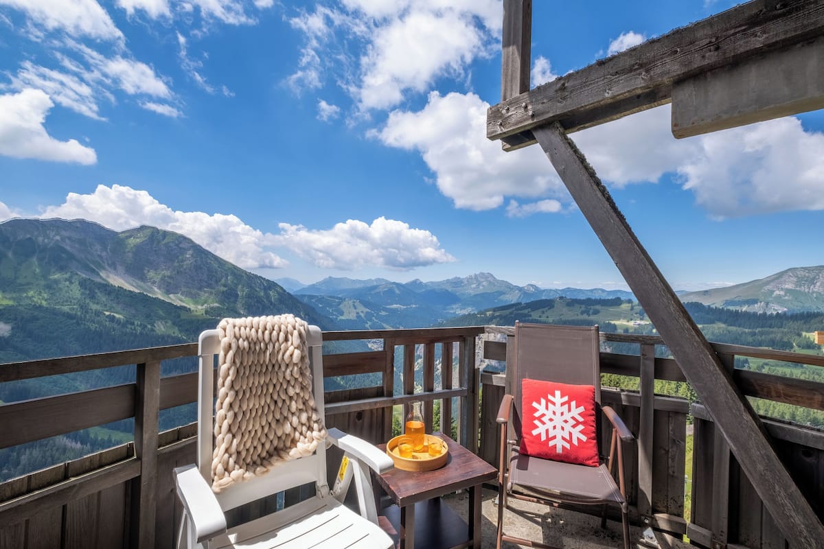 Eine Holzterrasse mit zwei Stühlen, einer mit weißem Kissen und der andere mit rotem Kissen, mit Blick auf eine Berglandschaft.