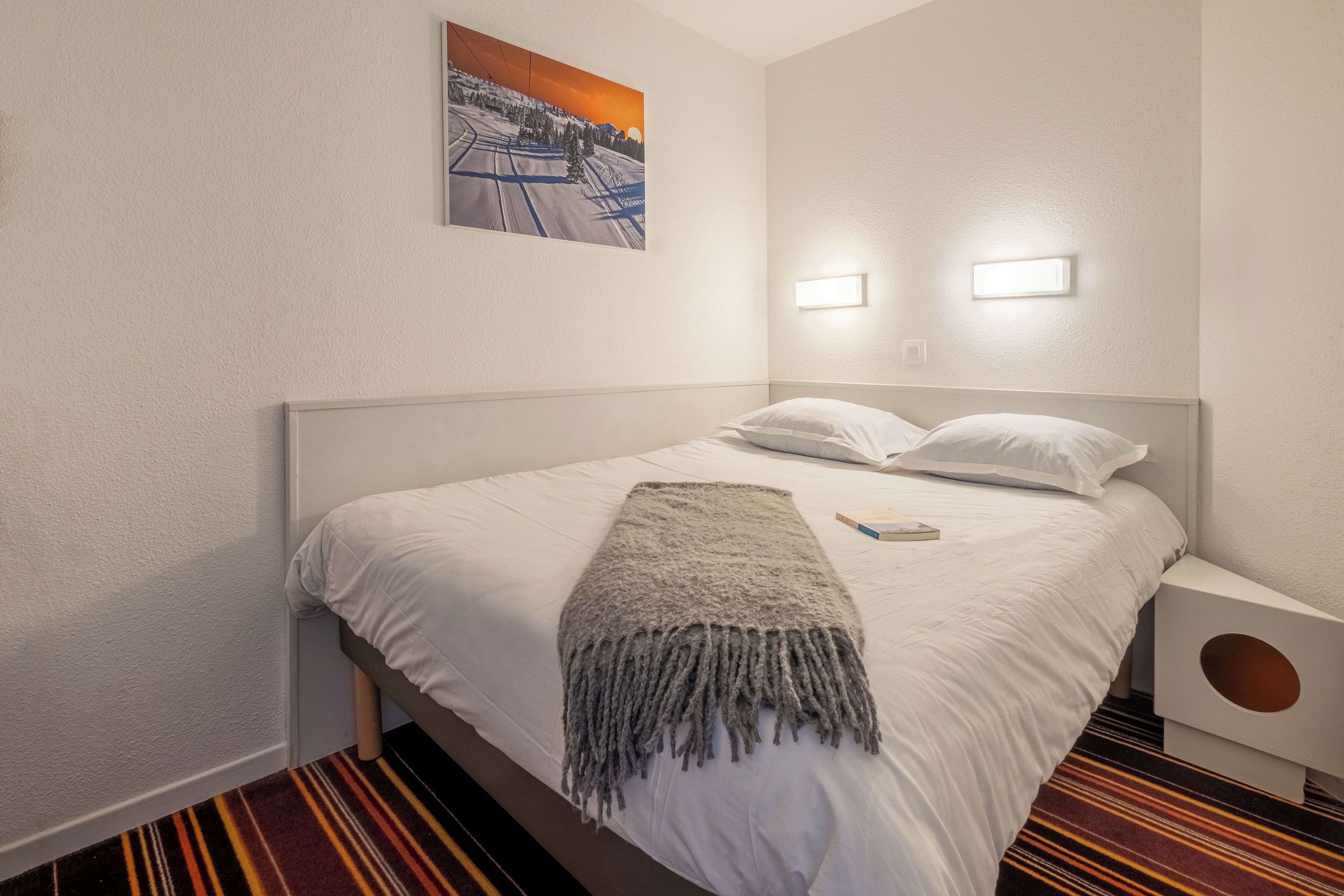 A neatly made bed with a white comforter and a gray throw blanket, a wall-mounted picture, and two bedside lamps.