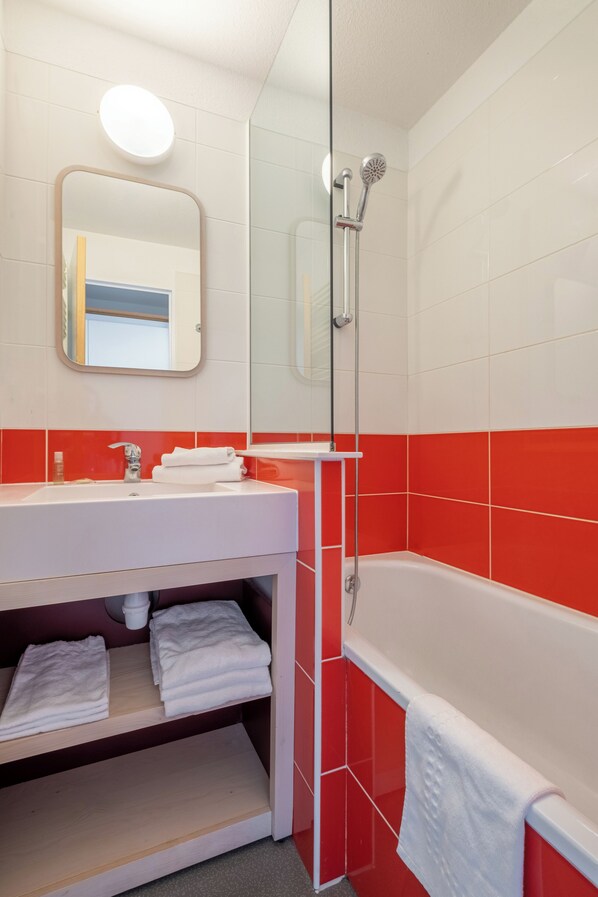 Un baño con lavamanos blanco, paredes de azulejos rojos, cabina de ducha con mampara de vidrio y espejo sobre el lavamanos.
