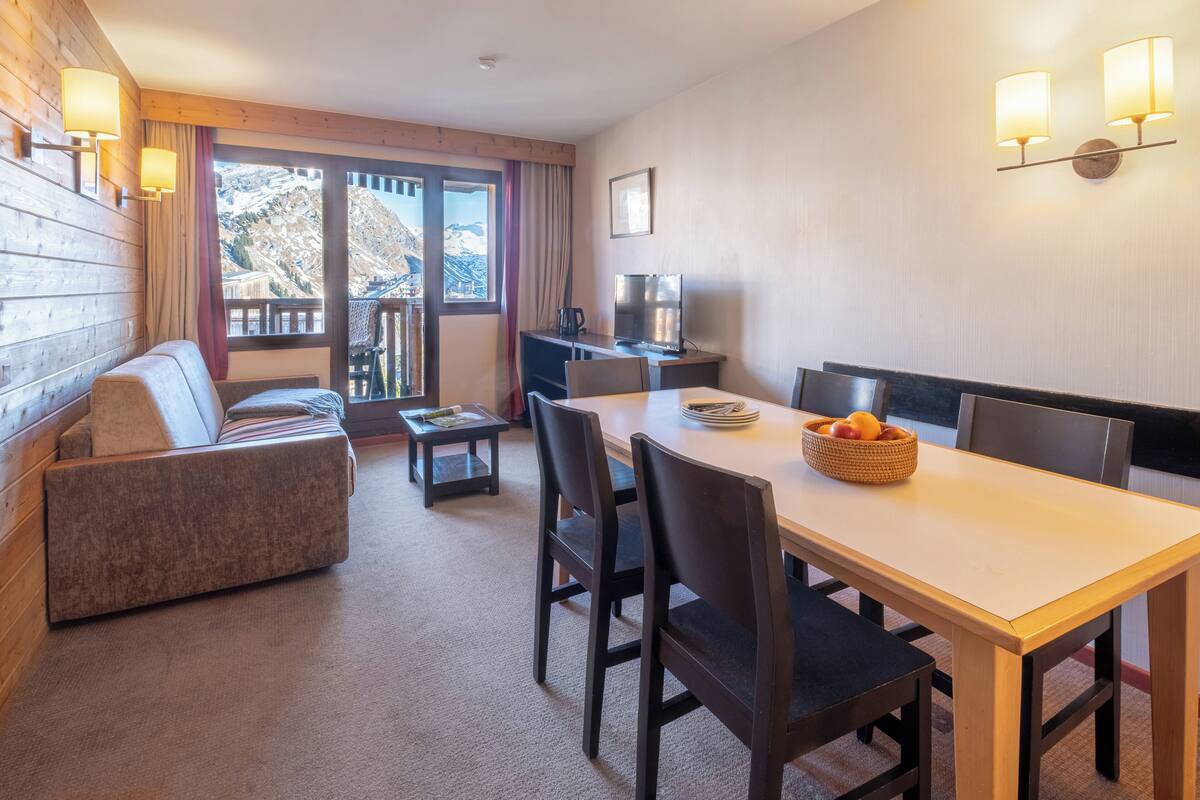 Ein gemütliches Zimmer mit einer Couch, einem Esstisch mit Stühlen, einem Fernseher und einem Fenster mit Bergblick.