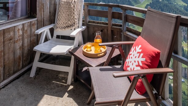 Ein Holzbalkon mit zwei Stühlen, einer mit rotem Kissen und der andere mit weißem Sitzkissen, mit Blick auf eine gebirgige Landschaft.