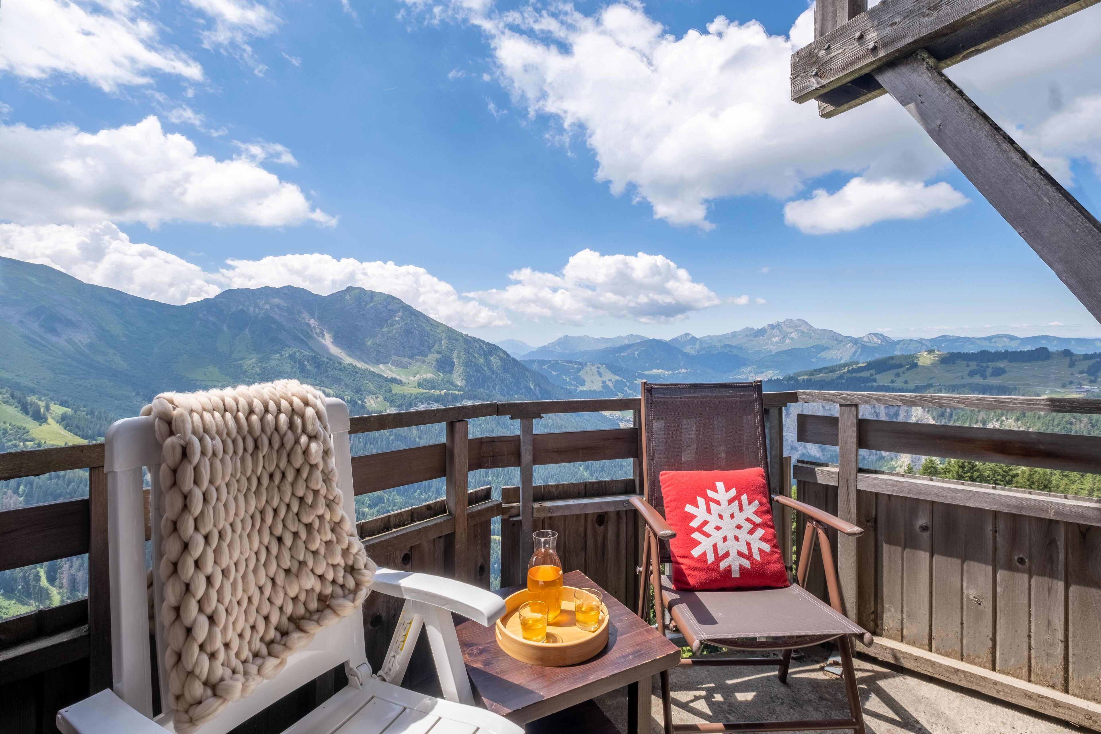 Une terrasse en bois avec une chaise et une table, une bouteille de boisson et un coussin rouge orné d’un motif en forme de flocon de neige.