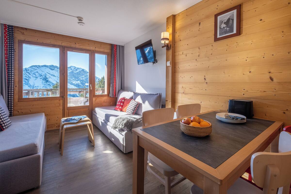 Ein gemütliches Zimmer mit einer Couch, einem Esstisch mit Obstkorb und Blick auf schneebedeckte Berge durch große Fenster.