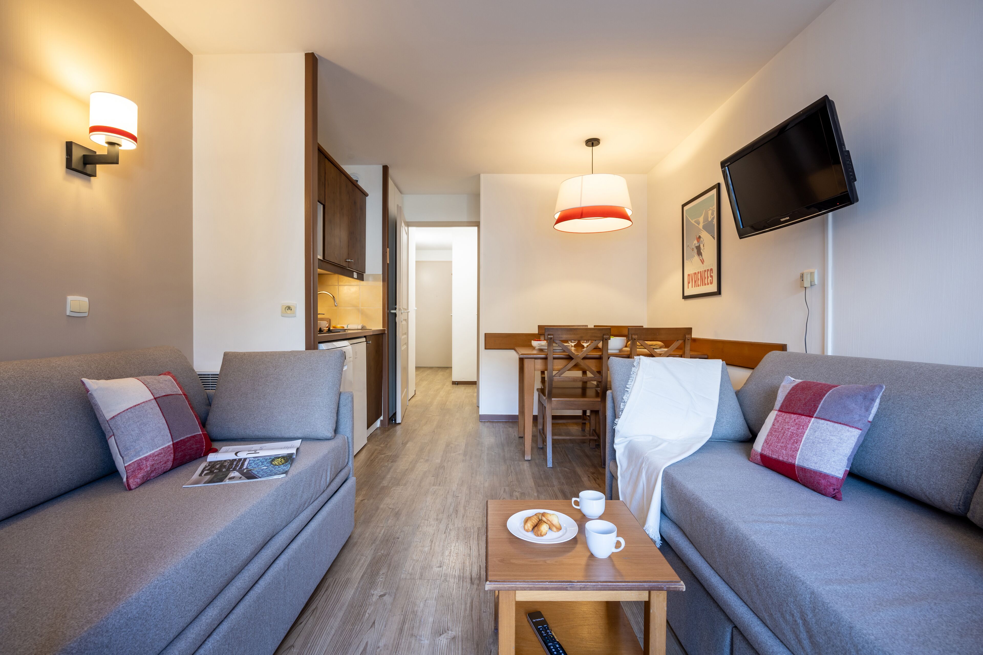 Una habitación de hotel moderna con dos sofás grises, una mesita de madera con un plato de pasteles y dos tazas, un televisor montado en la pared y una zona de comedor con una mesa y sillas de madera.