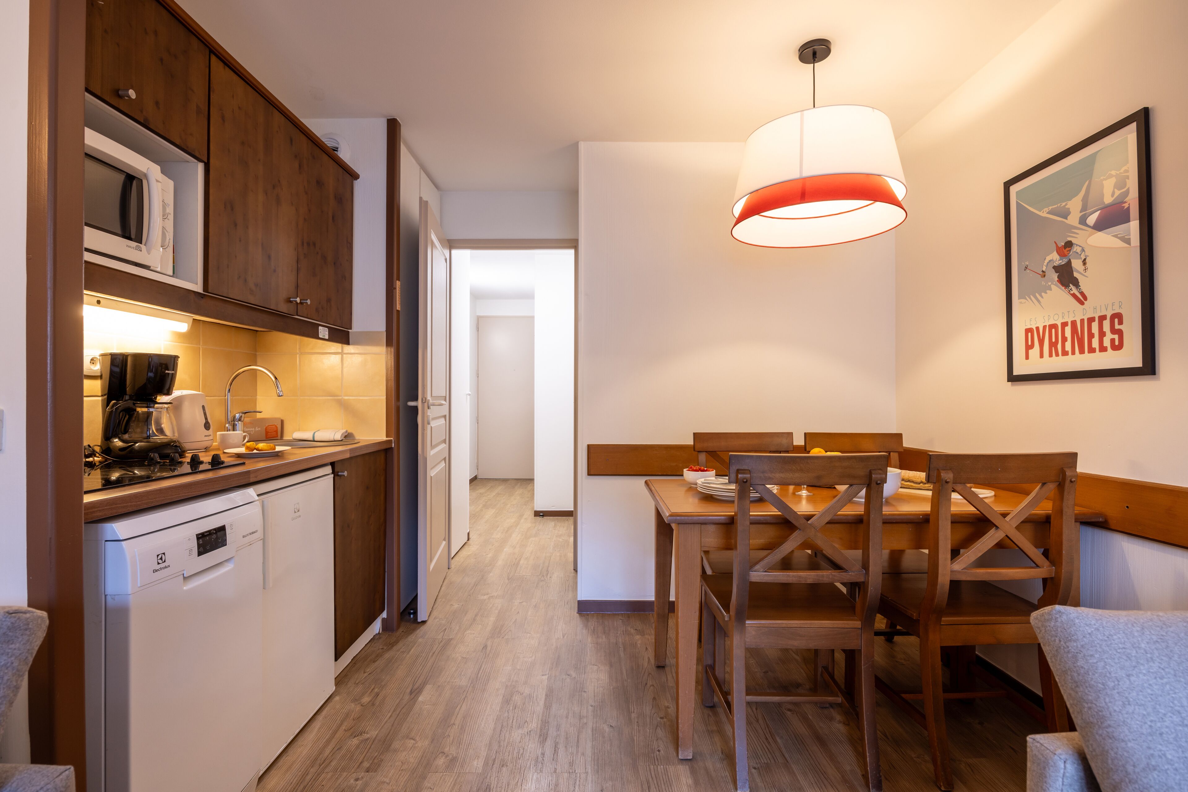 A compact kitchen with wooden cabinets, a microwave, and a dishwasher. There's a dining area with a wooden table and chairs, and a pendant light above.