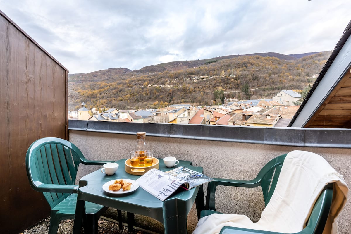 Een balkon met een ontbijtje, inclusief croissants, een fles sap en een krant. Op de achtergrond is een stad met gebouwen en heuvels te zien.