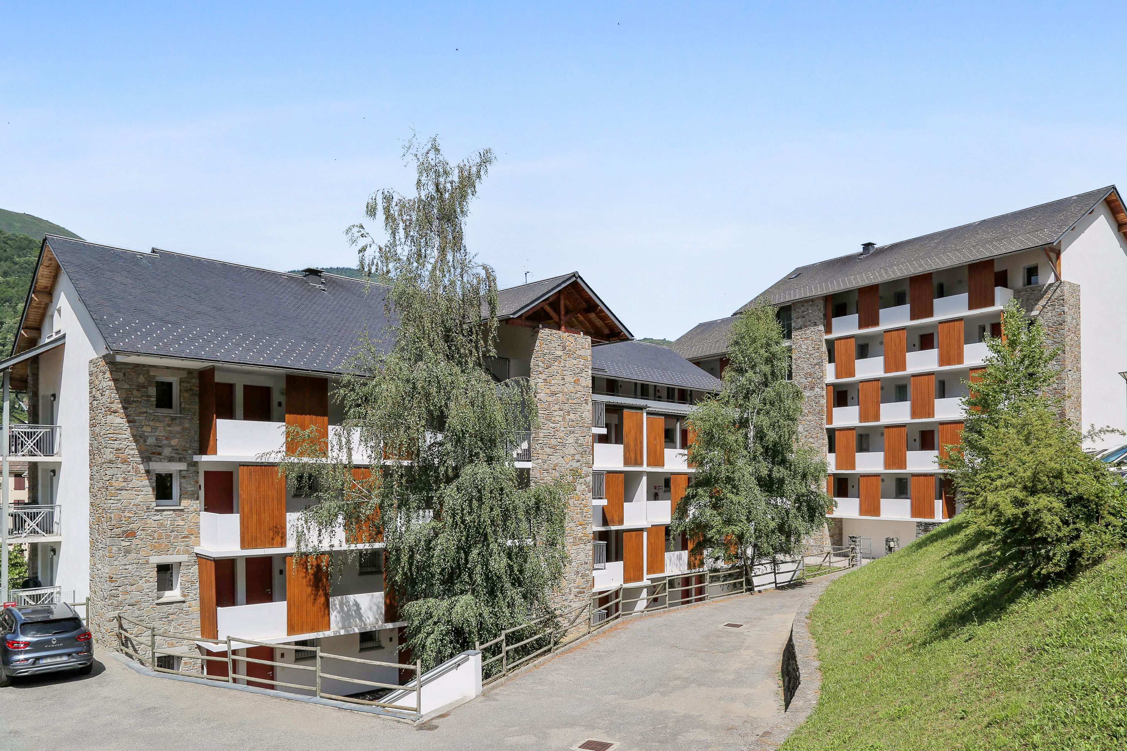 A modern multi-story building with balconies, stone and wood accents, and a parking area.