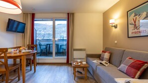 Habitación de hotel moderna con una mesa de comedor de madera, un sofá, un televisor y un balcón con vistas a las montañas.