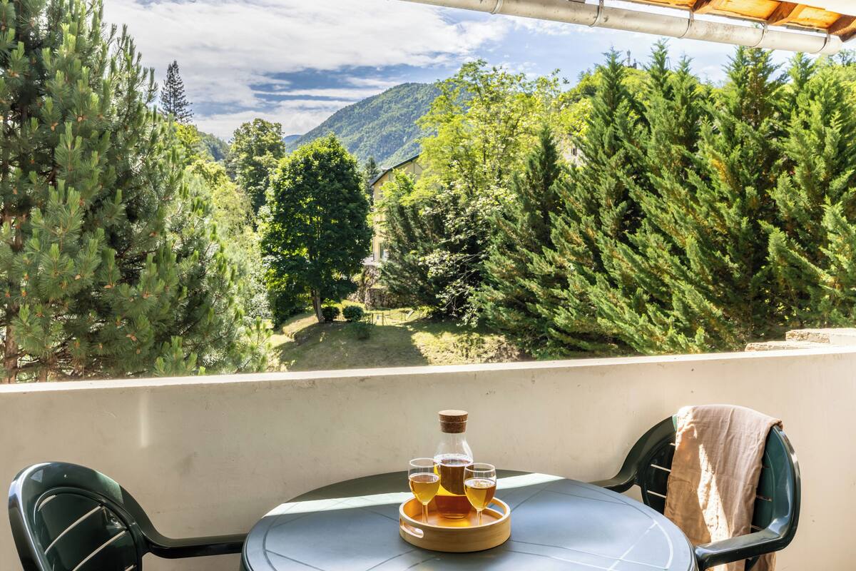 Een balkon met een tafel gedekt voor twee personen, een fles en twee glazen met een drankje. Op de achtergrond een bergachtig landschap en weelderige begroeiing.