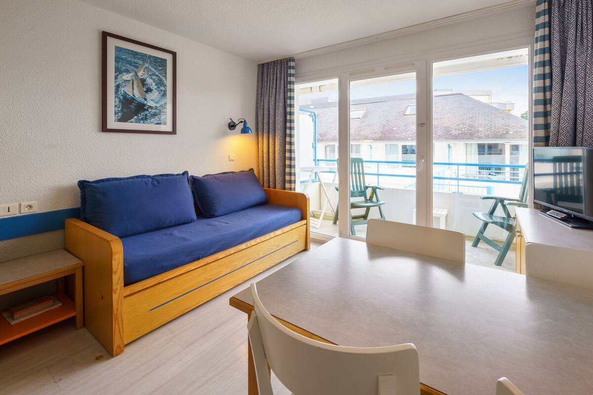 Ein Zimmer mit einer Couch, einem Tisch, Stühlen, einem Fernseher und einem Balkon mit Geländer.