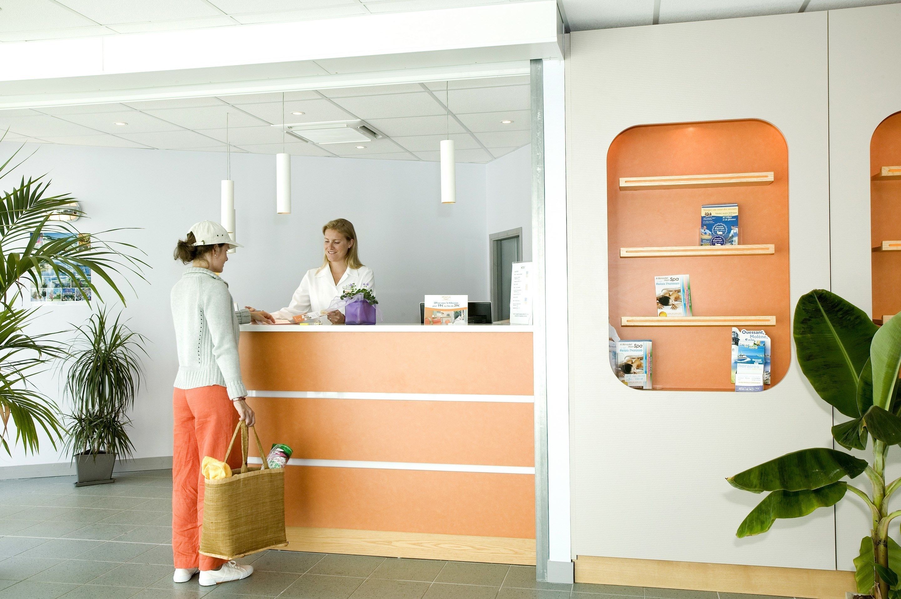 Un espace d’accueil comprenant un comptoir, une réceptionniste et un visiteur. Des plantes et des brochures d’information sont disposées sur des étagères.