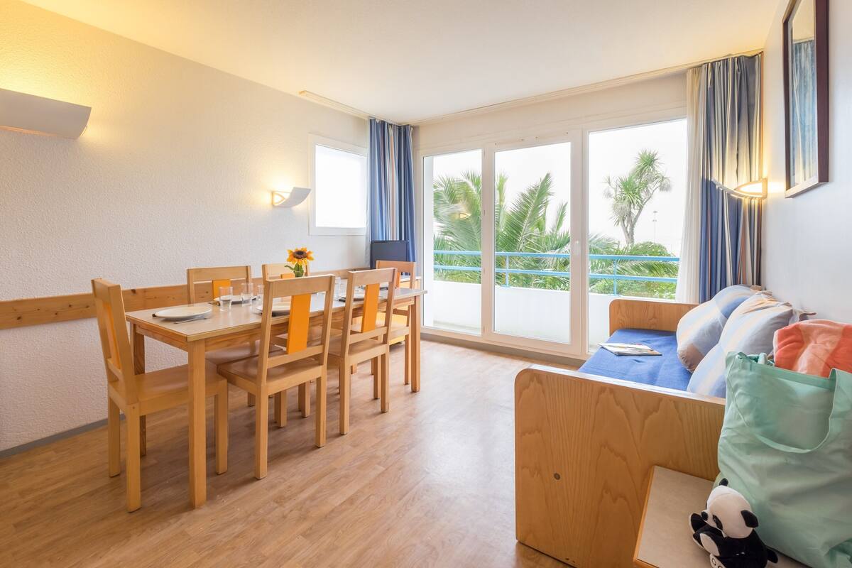 Ein Zimmer mit einem Holztisch und Stühlen, einem Sofa mit blauem Kissen und einem Fenster mit Vorhängen.