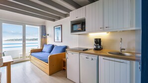A compact kitchen with a built-in microwave, a sink, and a dishwasher. There is a wooden bench with blue cushions in the living area, and a view of the sea through large windows.
