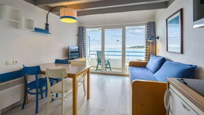 A room with a dining table, chairs, a sofa, a TV, and a view of the sea through large windows.