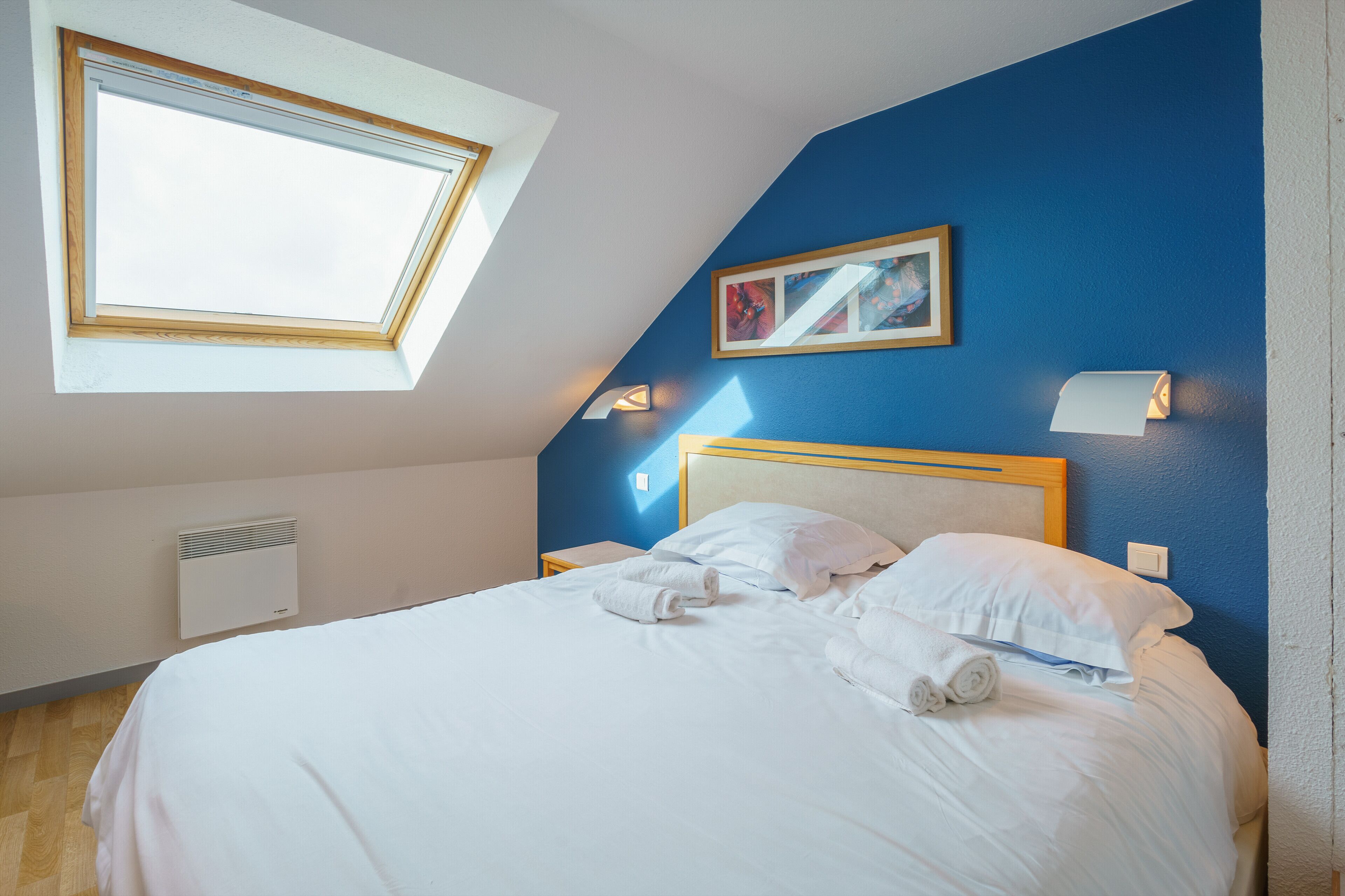 A neatly made bed with white linens and towels, a skylight window, a framed picture on the wall, and a bedside lamp.