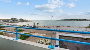 Uitzicht op de kust vanaf een balkon met een balustrade, een weg met geparkeerde auto's, een voetpad en een wateroppervlak.